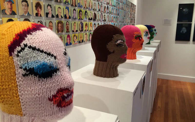 Knitted faces with portraits hanging on the wall behind them