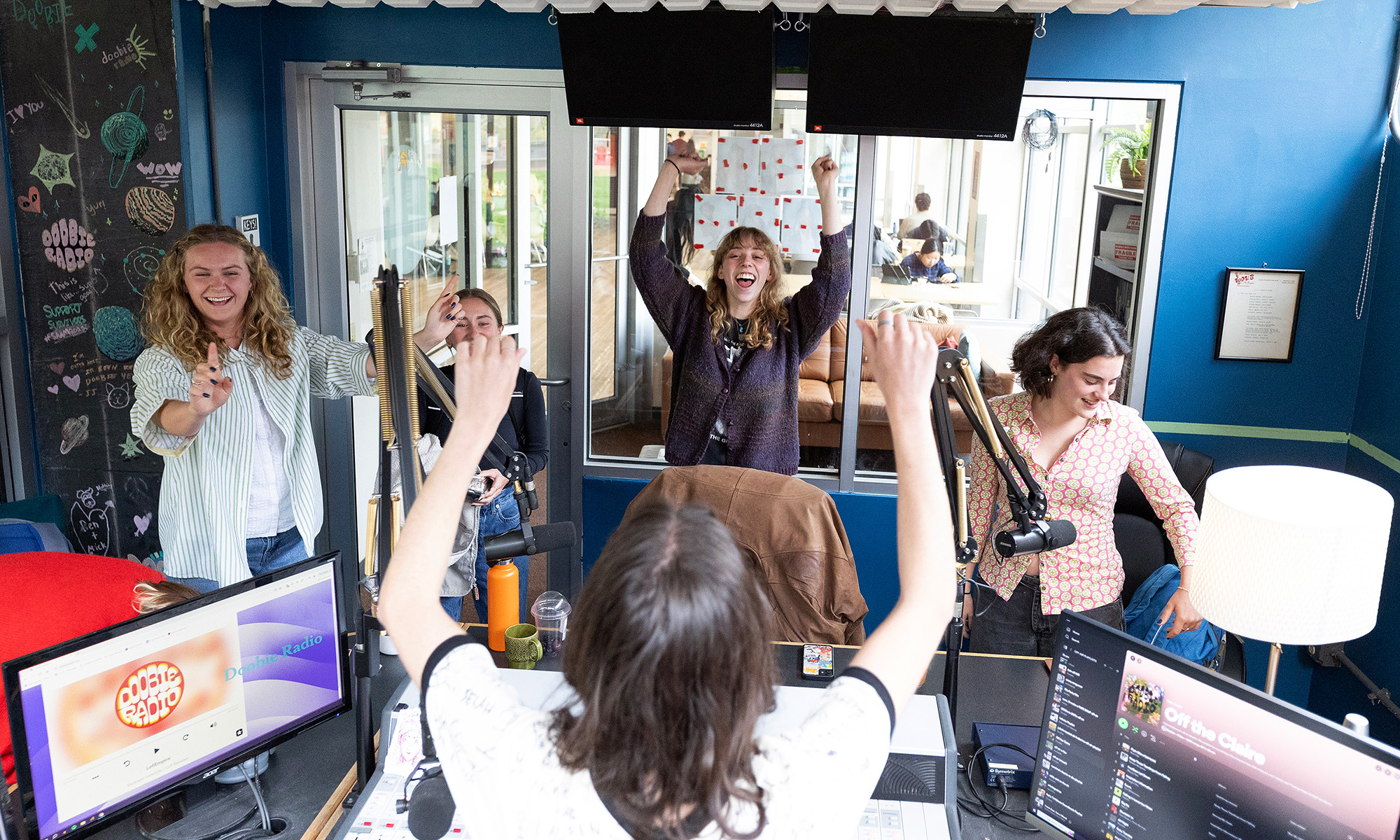 Five people in and around a radio broadcasting booth with their arms raised in cheers. 