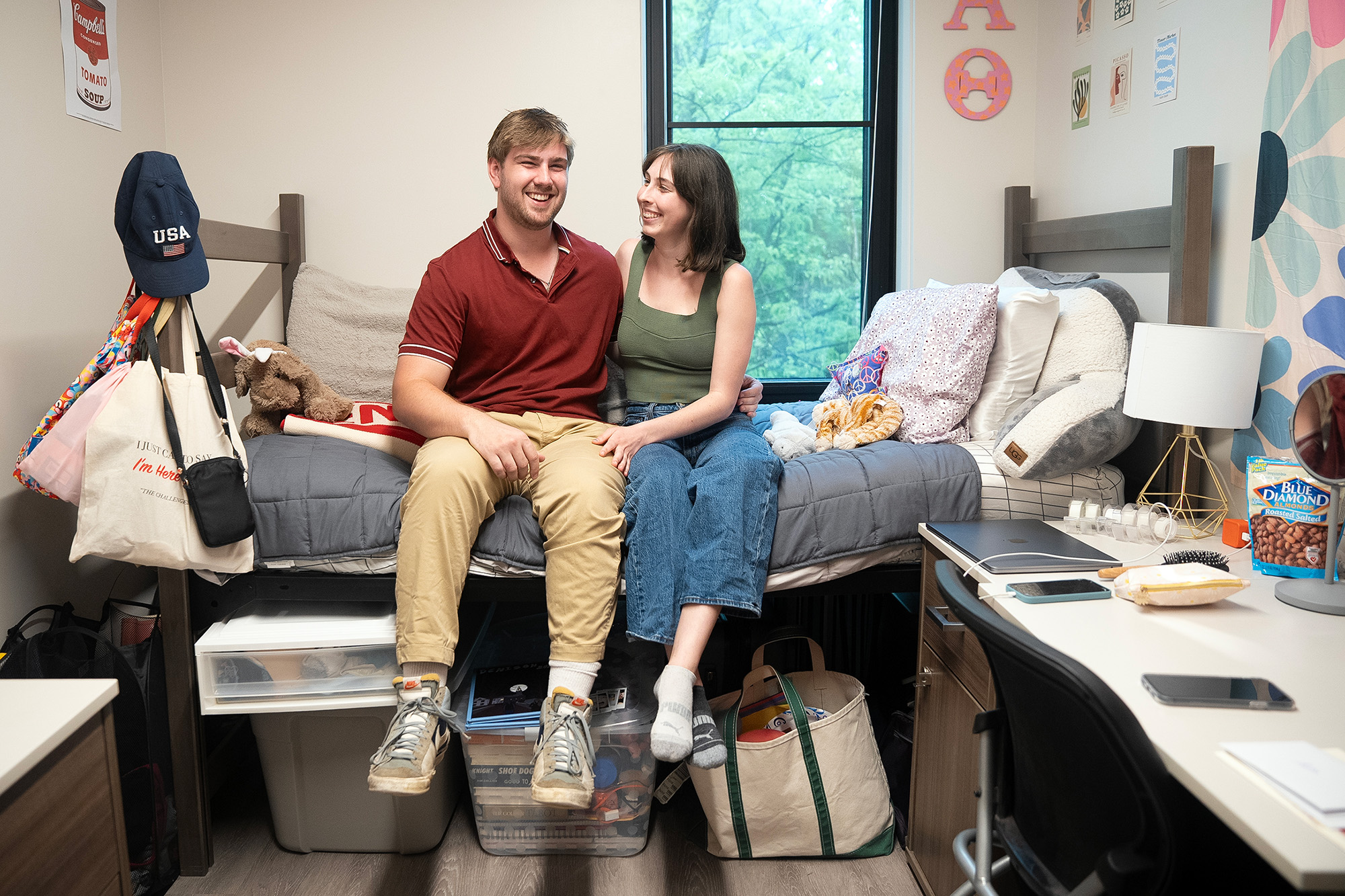 A man and woman sitting on a lofted bed in a college residence hall. The woman is looking at the man. Both are smiling. 