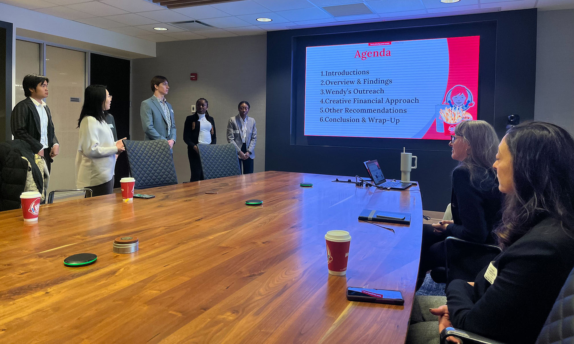 Photo of a conference room. Standing on the left side of the photo are four Denison students in business attire presenting a project, which is displayed on a wall screen (pictured center); at right, their audience, Wendy's President, International and Chief Development Officer Abigail Pringle ’96, and Rosa Ailabouni, a professional consultant who mentored the students during the semester-long project. 