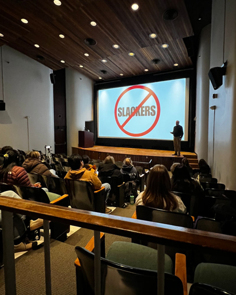 Photo of Rick Coplin ’85 giving a presentation to an auditorium full of students. The slide on screen displays the word "slackers" with a large red circle around it and a red diagonal line crossing through the word.