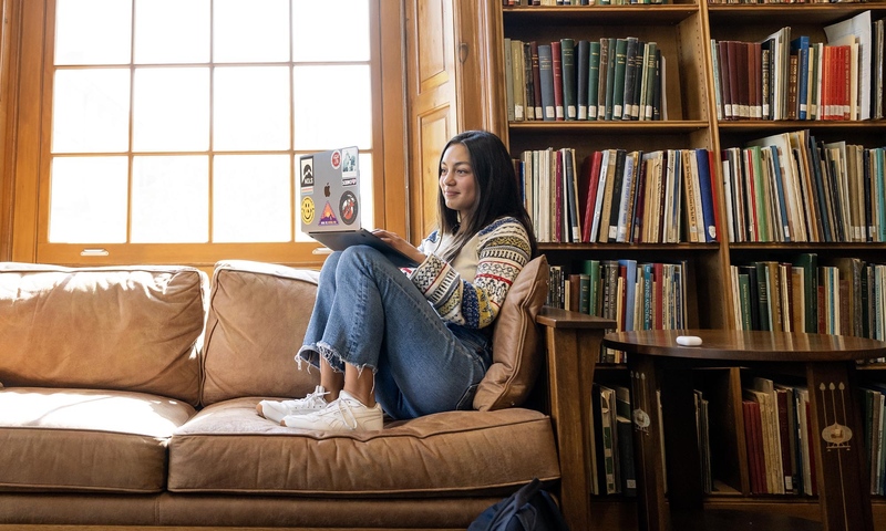 Student sitting on a sofa with a bright window and full bookcase behind her, holding a laptop computer on her lap.