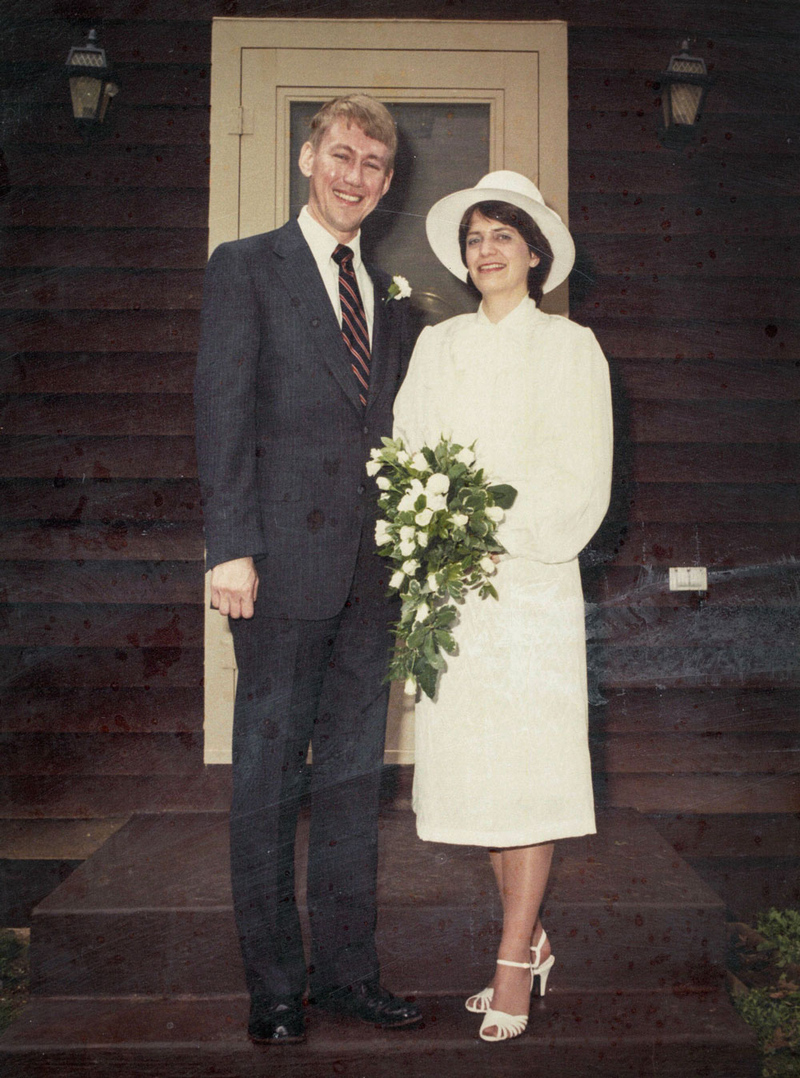 Man and woman stand beside one another outside of a door. He's in a suit; she's wearing a white dress and holding a bouquet of flowers.