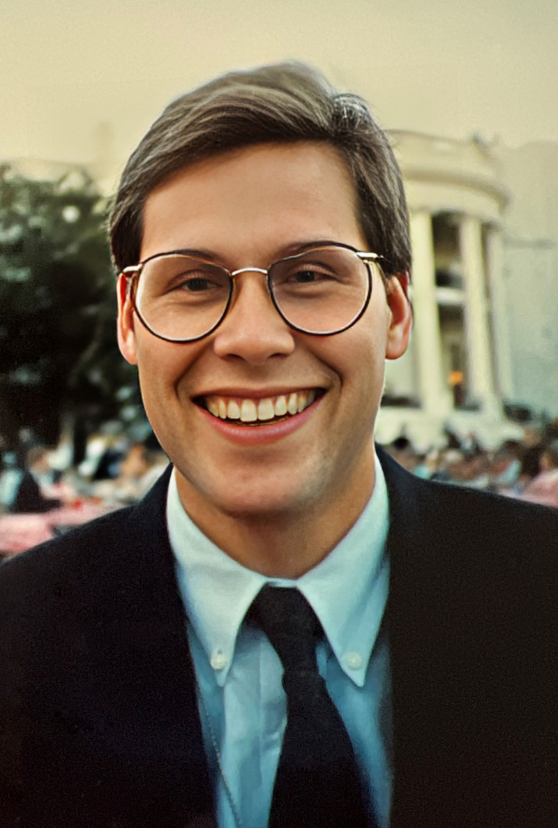 Man smiling, wearing glasses and a neck tie. The White House is in the background.