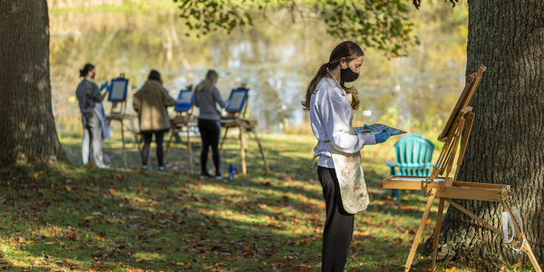 Students paint on easels outdoors while wearing masks