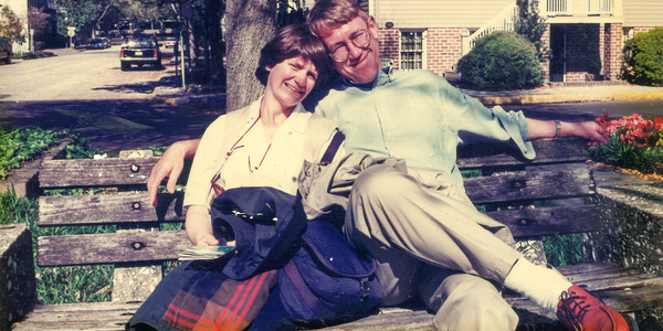 A man and a woman sitting on a park bench on a warm day. His arm is around her and she is leaning into him.