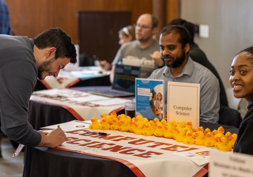 A student signs the Computer Science banner on a table lined with rubber ducks. There is a professor and a student sitting behind the table.