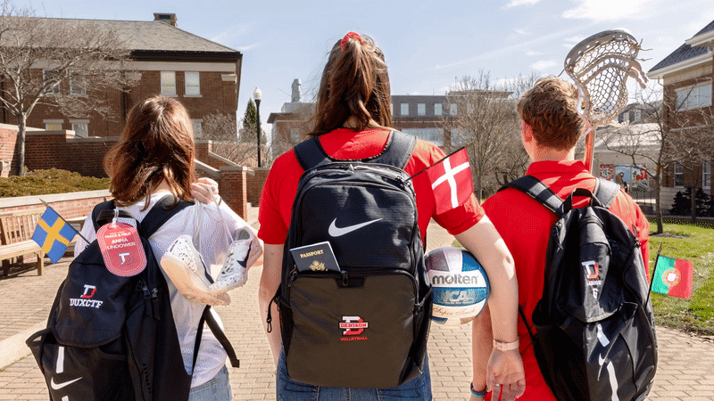 -Three athletes walking across the quad holding athletic gear. Their backpacks have flags sticking out of it that move slightly with the wind.